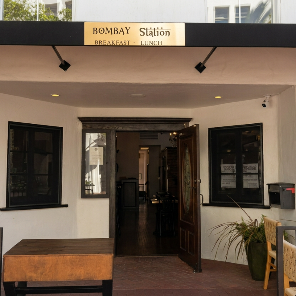 Exterior entrance of Bombay Station restaurant, with breakfast and lunch signage, outdoor tables, chairs, and potted plants.